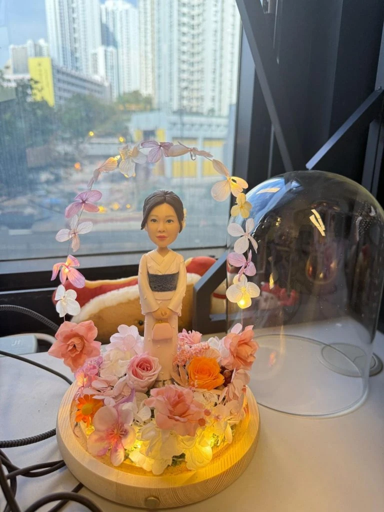 穿和服的人像公仔站在木座上,周圍以粉橘與米白永生花環繞,頭頂有花瓣燈串形成拱門,底部嵌入暖黃燈,旁放透明玻璃罩