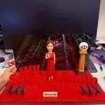 舞台式客製公仔擺設：女子穿紅色長禮服站在紅毯底座中央，背板是夜景霓虹與銀行大樓；台面立有「INV Sales Team」紅字樣、兩側麥克風架與HSBC標誌獎座，前方名牌寫著「Winnie Ng」。