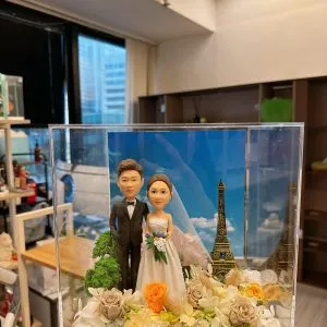 一對婚禮人像公仔站在花藝佈置的草地上,身穿黑色禮服與白色婚紗手捧花束,後方是藍天白雲與迷你巴黎鐵塔元素,整體置於透明展示盒內並隱藏暖黃小燈