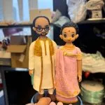 一對情侶公仔並肩站立：男子穿白色長袍配金紋披肩與眼鏡；女子著粉紅色紗麗加金色刺繡、額點紅痣與鼻環；腳下是藍黑圓台前緣擺放粉色愛心與玫瑰小飾。