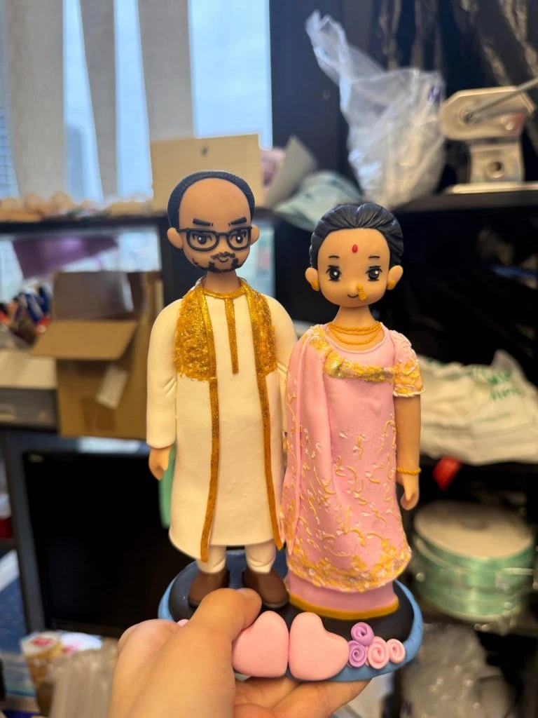 一對情侶公仔並肩站立：男子穿白色長袍配金紋披肩與眼鏡；女子著粉紅色紗麗加金色刺繡、額點紅痣與鼻環；腳下是藍黑圓台前緣擺放粉色愛心與玫瑰小飾。