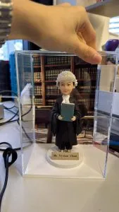 穿黑色法袍、戴傳統大律師假髮的女律師人像公仔,手捧綠色法律書站在名牌底座上,置於透明展示盒內,背後是書櫃背景