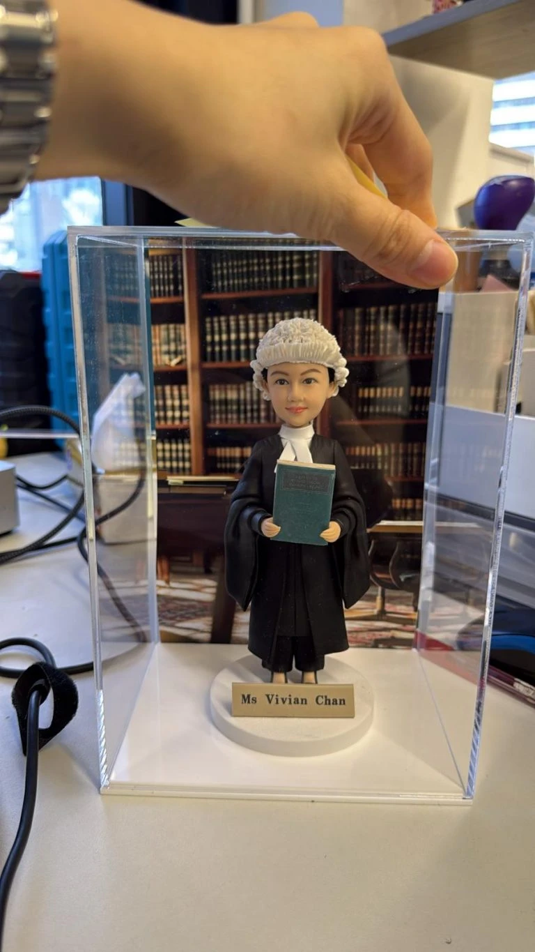 穿黑色法袍、戴傳統大律師假髮的女律師人像公仔,手捧綠色法律書站在名牌底座上,置於透明展示盒內,背後是書櫃背景