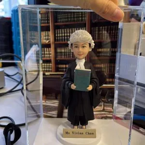 女大律師人像公仔戴傳統白色法庭假髮與黑色法袍，手持綠色法律手冊，置於透明展示盒內，前方金色銘牌刻 Ms Vivian Chan，背景為書牆律所場景