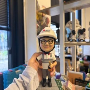 工程師主題人像公仔戴白色安全帽與對講耳機，穿公司POLO制服站在工地磚紋底座，手持香檳並配有姓名銘牌 Franklin Sare