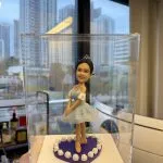 透明展示盒內的芭蕾公仔：女孩戴小皇冠，穿淡藍色薄紗舞裙與芭蕾舞鞋，雙手置前微屈膝擺出優雅姿勢；腳下為紫色愛心底座，邊緣圍繞白色玫瑰裝飾。