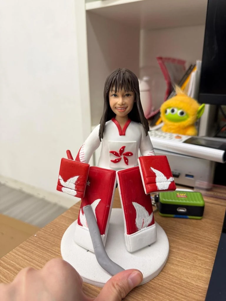 冰球門將主題公仔：長髮女孩穿白色球衣配紅色護具，胸前為五瓣花徽；雙手戴大面積護手、腿上穿紅白護腿，站在白色圓底座上微笑。
