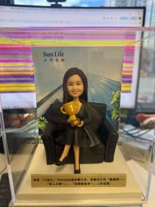 穿黑色套裝的女業務員人像公仔坐在黑色梳化上,手捧金色第一名獎盃,背後是公司大樓背景與 Sun Life 標誌