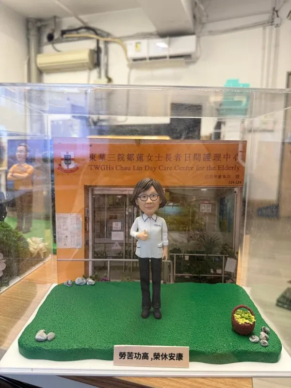 人像公仔穿白色制服站在綠色草地底座上，背後是「東華三院鄒蓮女士長者日間護理中心」門面背板，前方放有刻字銘牌，旁邊點綴小石與菜籃