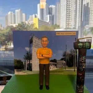 男士公仔穿橙色Polo衫與長褲,雙手抱胸站在綠色草地底座上,背後為機構建築背板,旁側擺放寫有「香港」字樣的圖騰路標,背板上貼有「平哥 Happy retirement 2025」