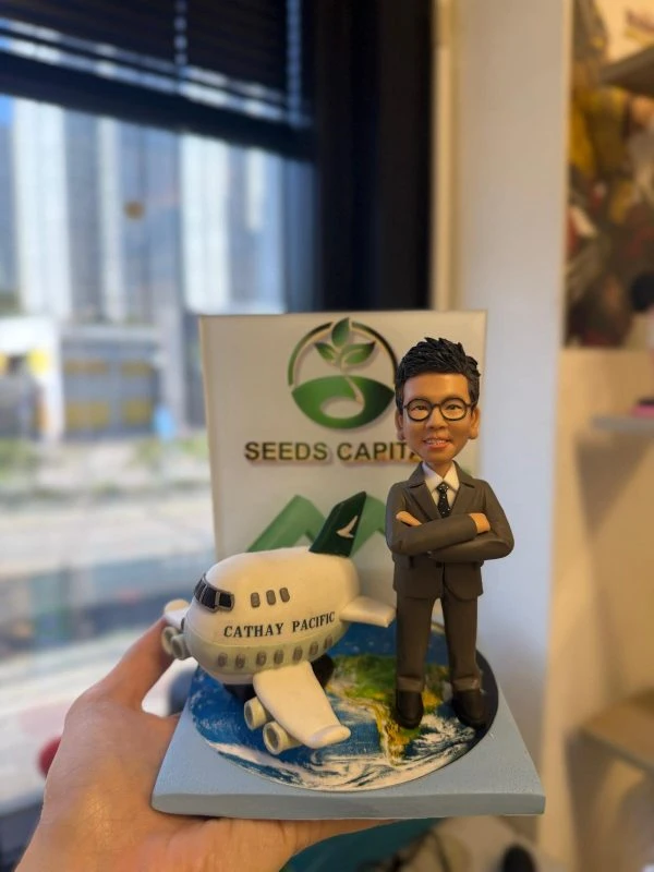 西裝人像公仔雙手交叉站在地球圖案底座旁，旁邊有印上 CATHAY PACIFIC 的小客機模型，後方立體背板寫有 SEEDS CAPITAL 與綠色標誌