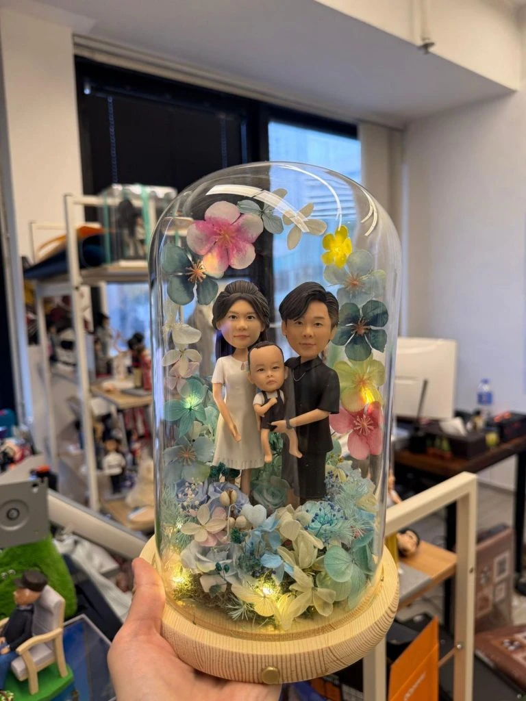 三人家庭人像公仔置於木座玻璃罩內，周圍以藍綠與粉色系花材環繞並點綴暖黃燈，呈現溫馨合照氛圍