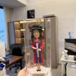 穿紅藍博士袍與博士帽的人像公仔站在透明展示盒內，手持卷起的畢業證書