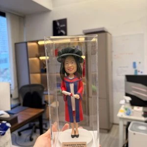 穿紅藍博士袍與博士帽的人像公仔站在透明展示盒內,手持卷起的畢業證書