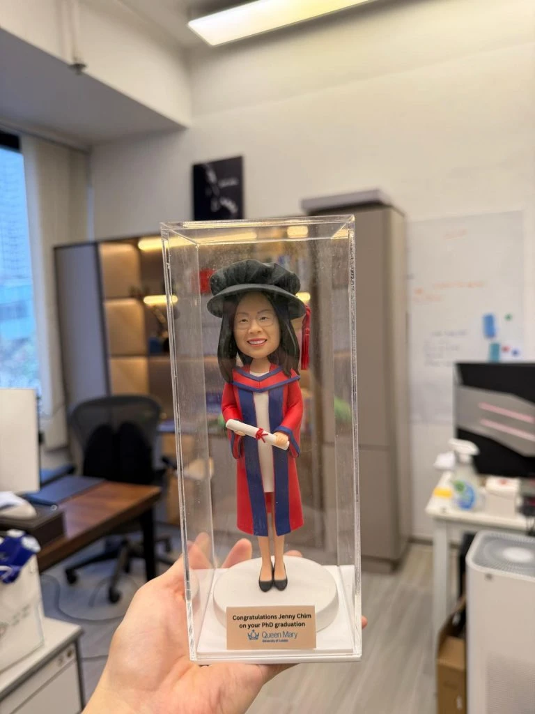 穿紅藍博士袍與博士帽的人像公仔站在透明展示盒內，手持卷起的畢業證書