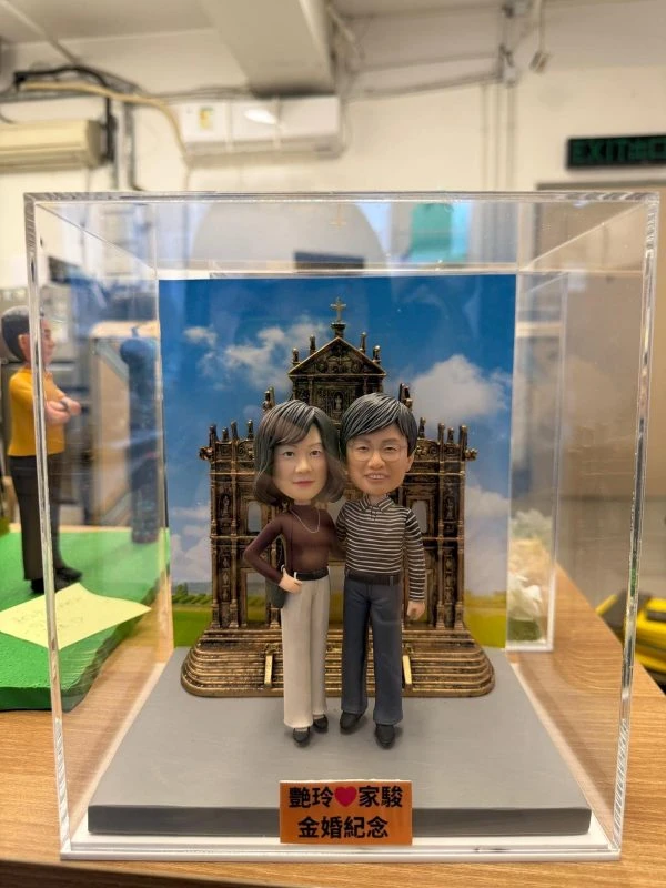 兩位人像公仔親密站立於灰色底座上，背後是澳門大三巴立體背板與藍天白雲圖，前方銘牌寫上金婚紀念字樣，整體置於透明展示盒內