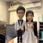 牽手站姿的婚禮公仔：男方戴眼鏡穿白色禮服外套配黑褲與黑色蝴蝶結，女方著蕾絲白紗與項鍊，立於白色圓形底座。