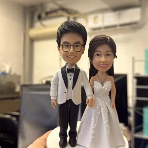 牽手站姿的婚禮公仔：男方戴眼鏡穿白色禮服外套配黑褲與黑色蝴蝶結，女方著蕾絲白紗與項鍊，立於白色圓形底座。