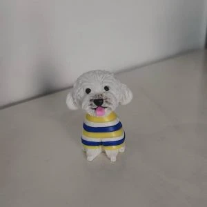 坐姿白色捲毛狗狗公仔,穿黃藍白條紋毛衣,黑鼻圓眼、粉紅舌頭微吐,放置在淺色桌面上。