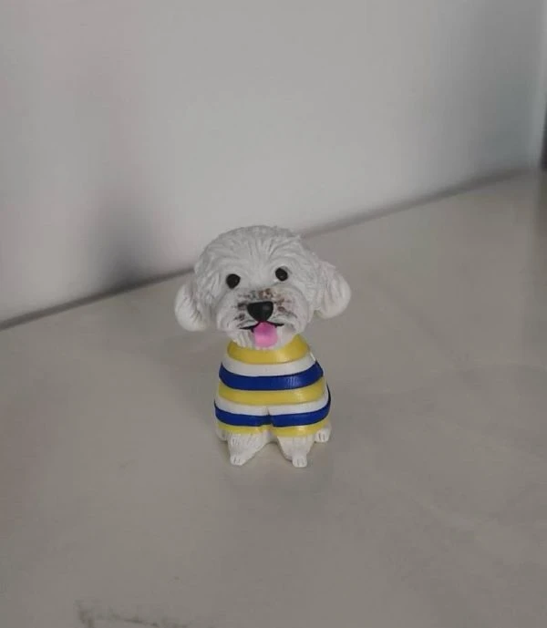 坐姿白色捲毛狗狗公仔，穿黃藍白條紋毛衣，黑鼻圓眼、粉紅舌頭微吐，放置在淺色桌面上。