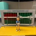 公仔坐在黃綠相間的排球門前，手抱排球，身穿藍色上衣與黑色短褲；背板為「香港仔消防局暨救護站」車房捲門，前方有致敬銘牌。