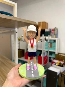 Alt: 女跑者公仔戴白帽掛金牌，雙手高舉站立於公路造型底座，馬拉松完賽紀念客製