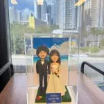 透明展示盒中的婚禮公仔：新郎著黑色西裝與蝴蝶結，新娘穿無肩白紗手捧粉橘花束，腳下綠草地台，背板為藍天白雲與海岸；前方小牌寫著「Keri & Tom September 20th, 2025」。