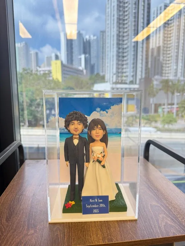 透明展示盒中的婚禮公仔：新郎著黑色西裝與蝴蝶結，新娘穿無肩白紗手捧粉橘花束，腳下綠草地台，背板為藍天白雲與海岸；前方小牌寫著「Keri & Tom September 20th, 2025」。