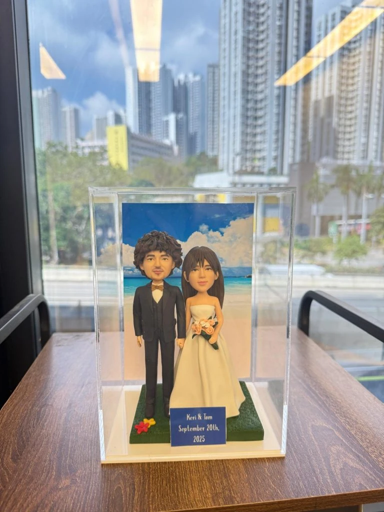 透明展示盒中的婚禮公仔：新郎著黑色西裝與蝴蝶結，新娘穿無肩白紗手捧粉橘花束，腳下綠草地台，背板為藍天白雲與海岸；前方小牌寫著「Keri & Tom September 20th, 2025」。