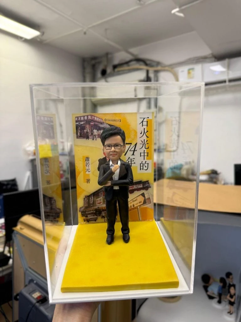 身穿西裝的手工軟陶單人公仔，置於透明展示盒內，背景為黃色書封場景，展現精緻雕塑與立體收藏效果。