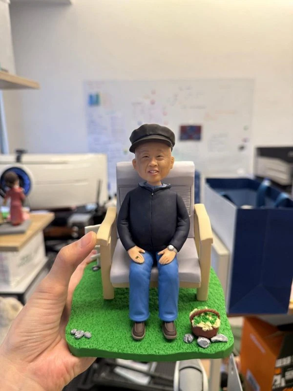 長者人像公仔戴報童帽、穿深色外套與藍褲，坐在木扶手椅上，置於綠色草地底座，旁邊有裝著蔬菜的小籃與石頭點綴，神情和藹