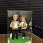 全手工製作的雙人軟陶公仔與一隻貓咪造型，置於透明展示盒與草地場景中，展現細緻神情與溫馨互動。