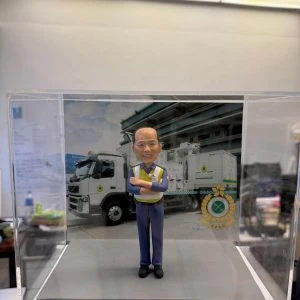 透明展示盒內的人像公仔:光頭男子穿藍色工作褲與反光安全背心,雙手抱臂站在灰色地台上;背板為工程車與作業場景,右下角有圓形徽章標誌。