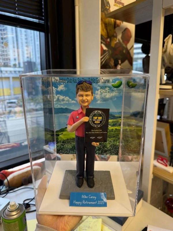 男士人像公仔穿紅色高爾夫上衣與黑長褲，手持「TOP 100 GOLF COURSE ASIA」獎牌站在六角磚地面上，背後是高爾夫球場風景背板，上有「The Jockey Club Kau Sai Chau Public Golf Course」字樣，前方藍色銘牌寫著 “Mike Carey Happy Retirement 2025”