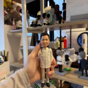 醫師主題人像公仔身穿白袍搭配聽診器,微笑比手勢站在灰色步道與綠地相間的小底座上