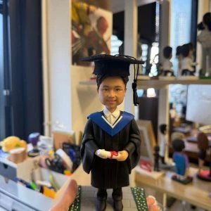 畢業主題人像公仔身穿黑色學袍與藍領披肩,頭戴學士帽,雙手捧著綁紅絲帶的畢業證書,站在兩側有小花點綴的灰色磚道底座上