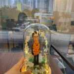 玻璃罩景觀公仔：男子抱著橘色毛茸茸小狗站在苔蘚草地上，周圍點綴藍白永生花與葉片形成拱門，底座為原木色並繞有暖黃燈串發光。