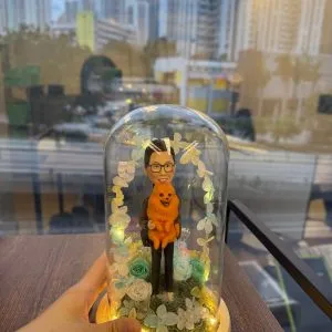 玻璃罩景觀公仔:男子抱著橘色毛茸茸小狗站在苔蘚草地上,周圍點綴藍白永生花與葉片形成拱門,底座為原木色並繞有暖黃燈串發光。