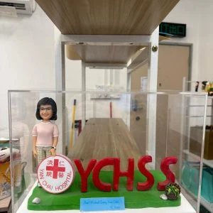 機構主題公仔搭配大型紅色 YCHSS 字母與院徽圓牌,草地底座置於透明展示盒,歡送致敬