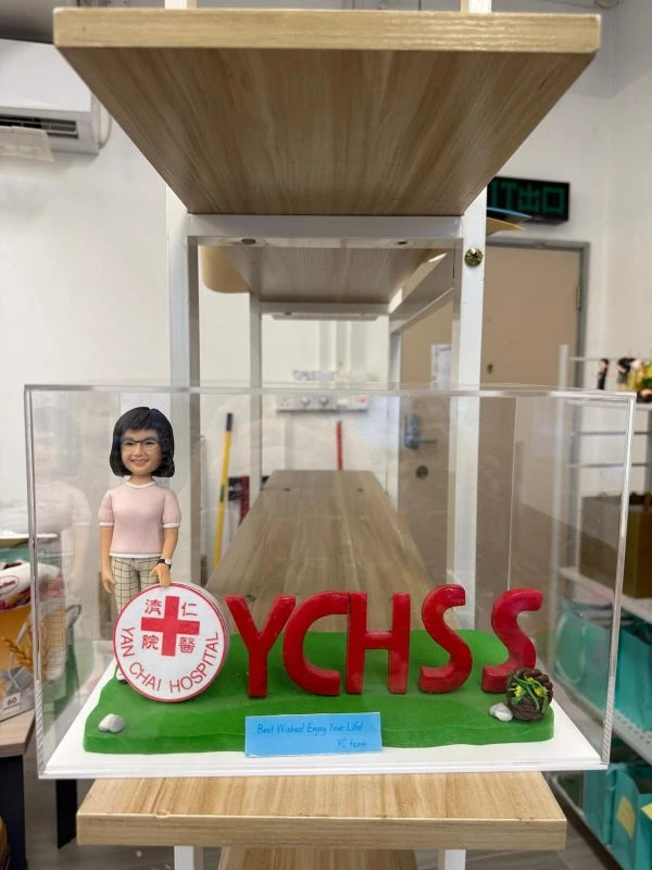 機構主題公仔搭配大型紅色 YCHSS 字母與院徽圓牌，草地底座置於透明展示盒，歡送致敬