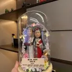 一對穿民族傳統禮服的夫妻人像公仔被裝在玻璃罩內，周圍環繞花環燈飾與木紋底座刻名牌