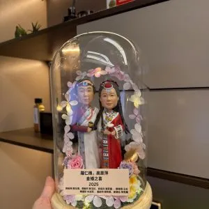 一對穿民族傳統禮服的夫妻人像公仔被裝在玻璃罩內,周圍環繞花環燈飾與木紋底座刻名牌
