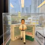 透明展示盒內的制服公仔：短髮男子穿白色襯衫與長褲、黑色皮帶與領帶，做雙手豎拇指手勢站在灰色磚道地台上；身後為綠白相間的病房指示牆與標誌，地台兩側有草皮花帶，前方金色銘牌寫有榮休祝福與科室名稱。