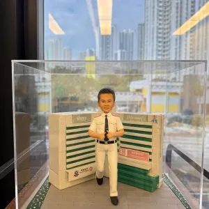 透明展示盒內的制服公仔:短髮男子穿白色襯衫與長褲、黑色皮帶與領帶,做雙手豎拇指手勢站在灰色磚道地台上;身後為綠白相間的病房指示牆與標誌,地台兩側有草皮花帶,前方金色銘牌寫有榮休祝福與科室名稱。