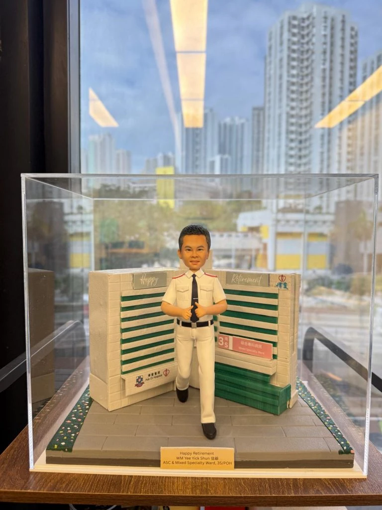 透明展示盒內的制服公仔：短髮男子穿白色襯衫與長褲、黑色皮帶與領帶，做雙手豎拇指手勢站在灰色磚道地台上；身後為綠白相間的病房指示牆與標誌，地台兩側有草皮花帶，前方金色銘牌寫有榮休祝福與科室名稱。