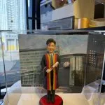 透明展示盒內的客製畢業公仔，男士穿黑色長袍配紅黃滾邊與領帶，手持捲軸站在紅黑底座上，背板為醫院大樓外觀，前方銘牌寫上致贈者訊息