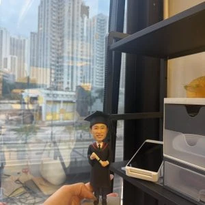 手持的客製畢業公仔,男生穿黑色學士袍與方帽、披紅邊披肩並拿著捲軸,底座銘牌寫「Happy Graduation Stephen 2025」