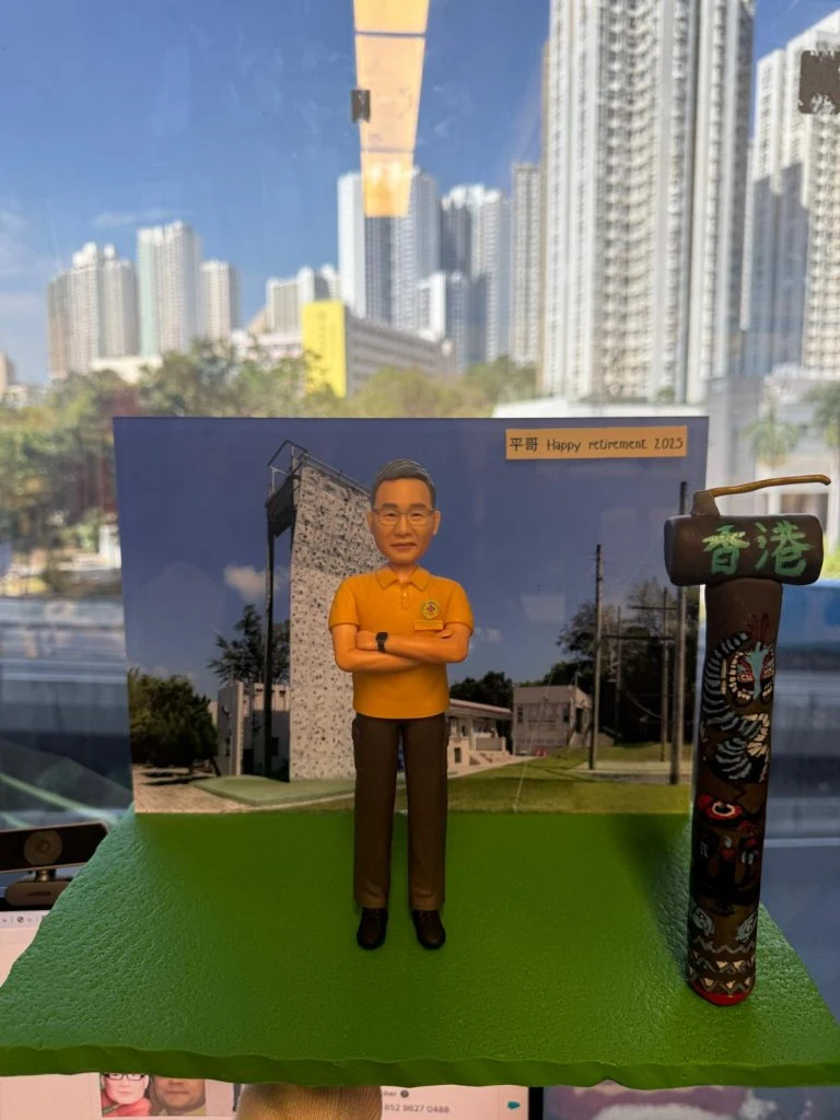 香港警隊致敬公仔成品；公仔穿橙色POLO與長褲、雙手交叉站立，背板為帶有攀岩牆紋理的建築場景，貼有「平哥 Happy retirement 2025」字條，右側擺放刻有「香港」字樣的裝飾柱