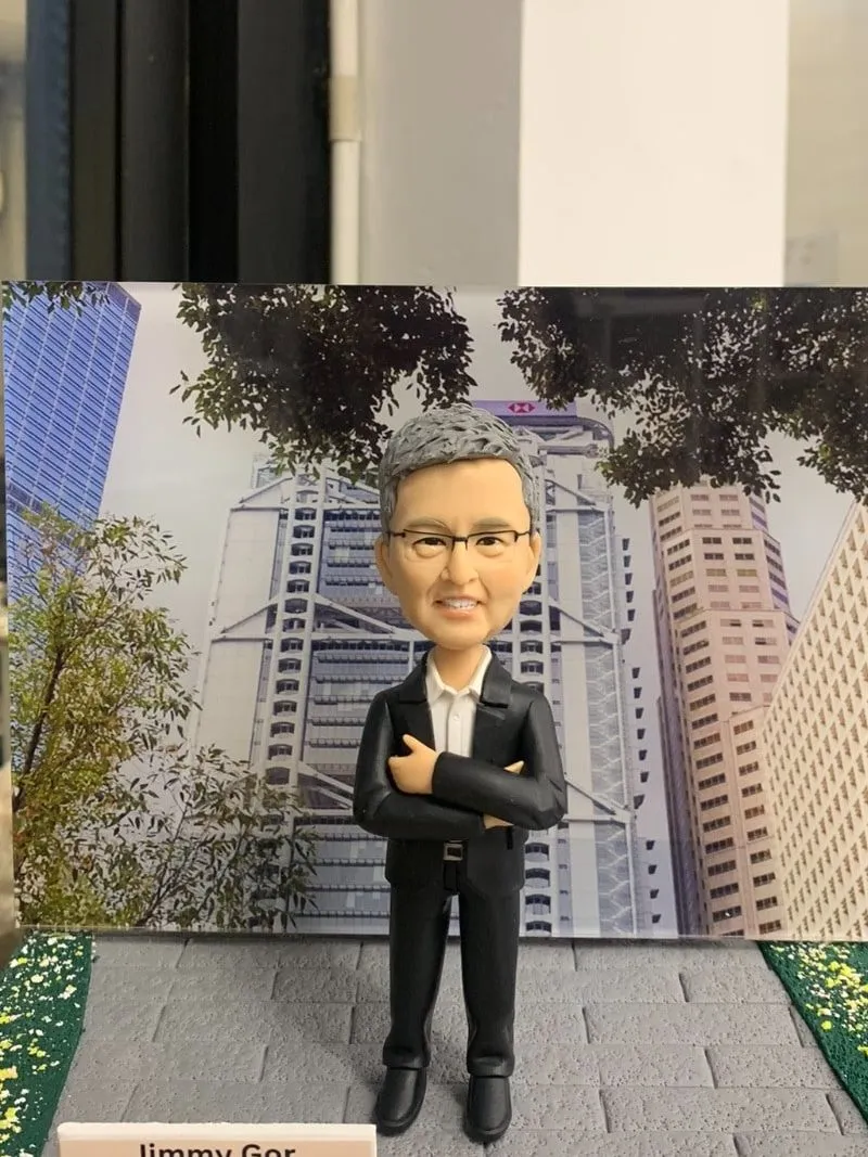 高層榮休主題的人像公仔：灰髮戴眼鏡的男士身穿黑色西裝與白襯衫，雙手抱胸自信站立於灰色磚紋地台中央；腳邊左前方立有白色名牌，上印「Jimmy Gor｜Happy Retirement」字樣。背板為都會金融區高樓與樹蔭交錯的城市景觀，象徵商務領域的職涯背景；地台兩側加入綠地灑花紋理，增添儀式感。整體以室內靠窗拍攝，光線清朗，呈現專業、尊榮且具紀念價值的企業高管人像公仔。