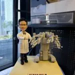 客製外科醫師公仔穿白袍打領結，伸手示意站在手術機器人旁，方形底座前有中英文銘牌